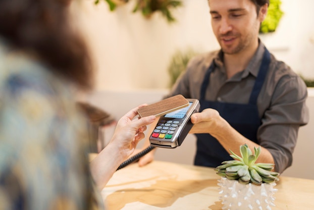 A male shop owner holds up a point-of-sale reader to a customer. The customer holds her smartphone to the reader to pay for her transaction.