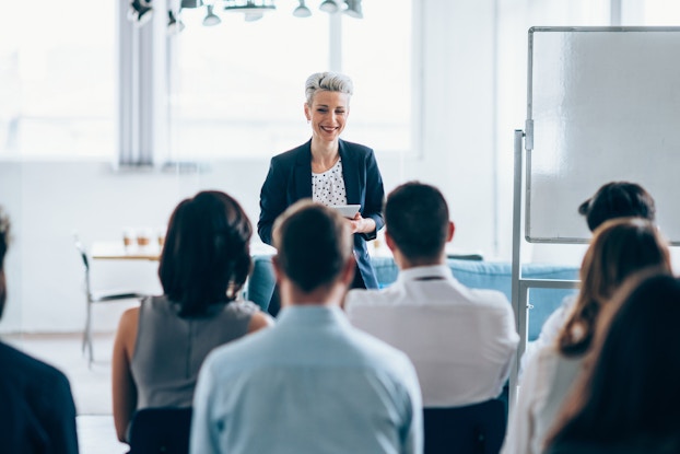A smiling woman with short silver-blonde hair stands at the front of a conference room, facing an audience of employees sitting in black plastic chairs. A blank whiteboard stands to the right of the woman.