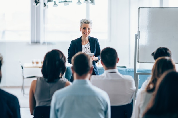  A smiling woman with short silver-blonde hair stands at the front of a conference room, facing an audience of employees sitting in black plastic chairs. A blank whiteboard stands to the right of the woman.