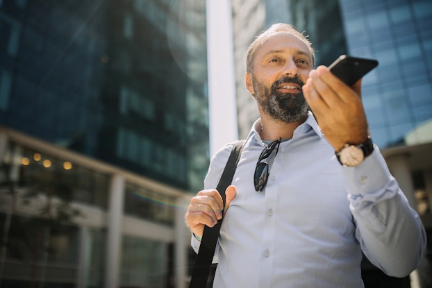  A man with a graying beard stands outside in front of a couple of mirrored skyscrapers. He wears a button-up shirt with a pair of sunglasses tucked into the collar and the strap of a bag over one shoulder. He holds a smartphone up near his mouth, ready to use it on speaker mode.