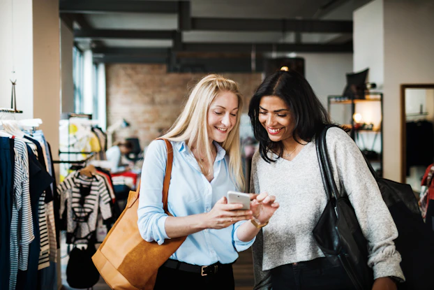 Two women stand in a clothing shop and look down at a smartphone. The woman on the left has long blonde hair and is wearing a light blue button-up shirt. She has a tan handbag over one shoulder. She is holding the smartphone and pointing at something on its screen. The woman on the right has long, dark hair and is wearing a pale gray sweater. She has a black handbag over one shoulder and she looks down at the smartphone with a smile.