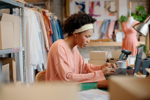 A woman in a back office of a clothing store sits at a desk, using a laptop. The woman has a concentrated look on her face as she types; she wears a light pink long-sleeved blouse and a yellow-and-white patterned headband. Directly behind her are freestanding metal shelves filled with boxes and a rack filled with hanging clothes.