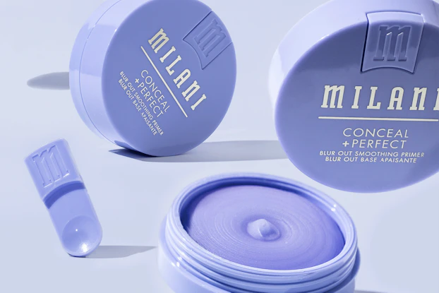Several jars of Milani Conceal + Perfect smoothing primer suspended at different angles so they look like they're floating. The jars are shallow periwinkle-colored cylinders with the Milani logo and product name printed in white on top. One of the jars is open; the primer cream inside is the same periwinkle color as the jars.