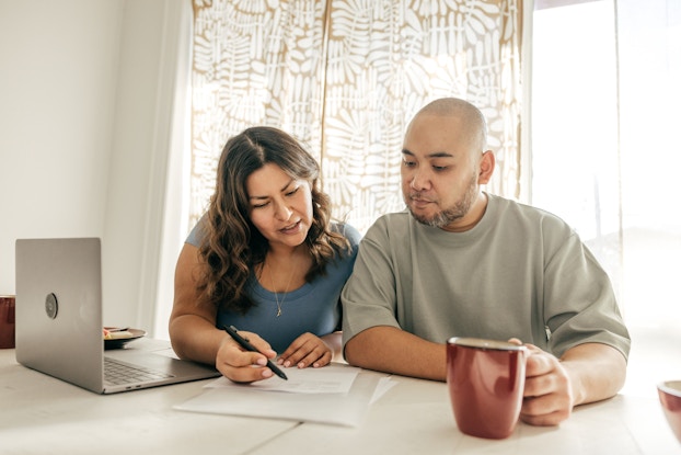 A couple sits at a table, looking at some papers in front of them. The person on the left, a woman with long, wavy dark hair, points to something on the paper with a pen. The person on the right, a man with a shaved head and graying beard, is holding a red coffee mug.