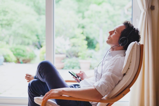 A man wearing headphones leans back in a chair with his eyes closed. The headphones are wired into a smartphone that the man holds in his right hand. In the background is a sliding glass door through which can be seen a patio, several potted plants, and a dense green forest.