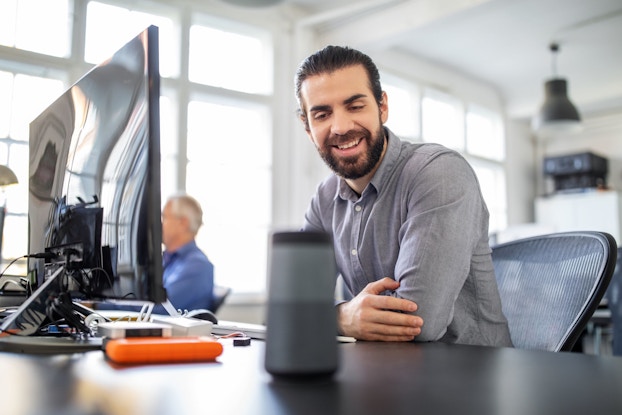 A man in a modern office is seated at a desk in front of a computer. He smiles as speaks to a digital speaker. He is asking a digital assistant a question.