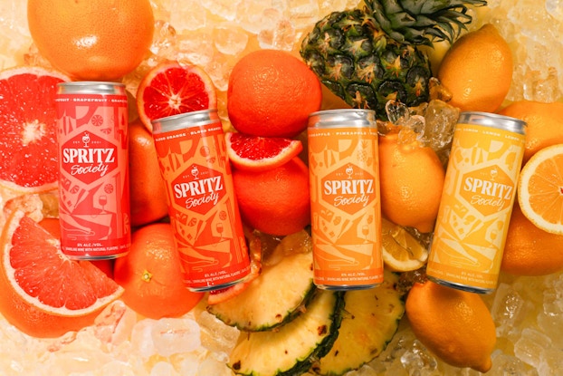  Four cans of Spritz Society canned wine cocktails displayed on top of fresh cut fruit.