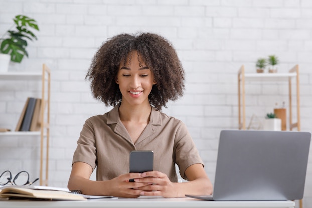  A smiling woman looks at her smartphone. An open laptop sits at her elbow.