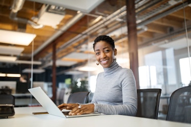 A woman looks at the camera and smiles. Before her is her laptop computer. Behind her is a modern open workspace.