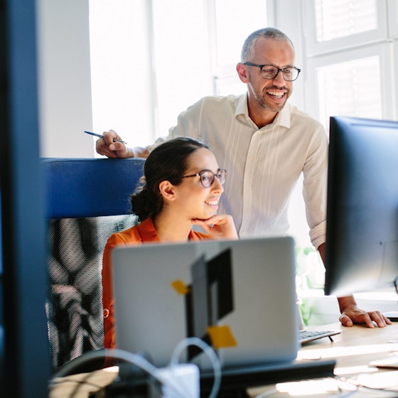A male and female work colleague are smiling while looking at a desktop computer monitor.