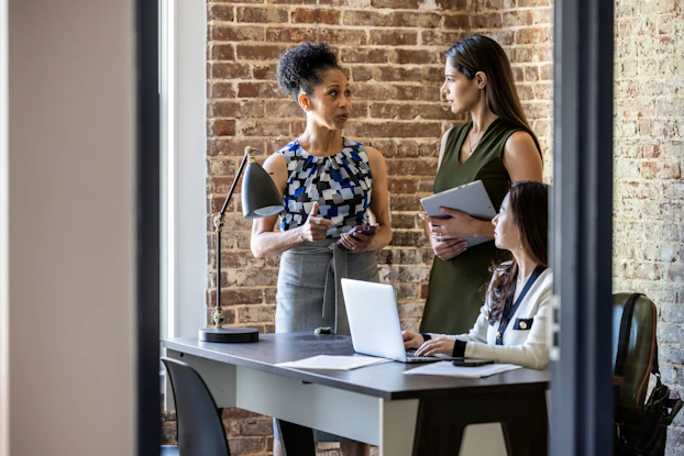 Three female colleagues at work are having a discussion. One of them is sitting at a desk.