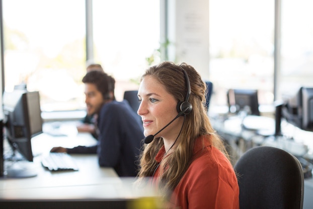  A smiling female customer representative wears headphones while talking to a customer. Behind her are other representatives who are on the phone with other customers.