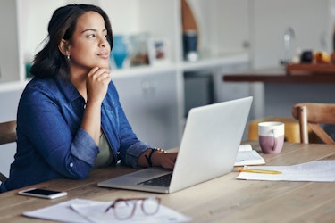 A young woman seated at a table in her kitchen thinks about something while using a laptop computer.