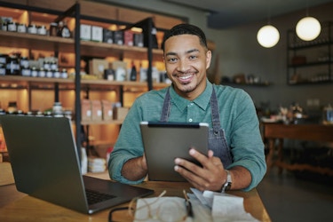 A shop owner smiles at the viewer. He is sitting at t able in his tore with his laptop open. He is holding a computer tablet and has his glasses and papers spread in front of him.