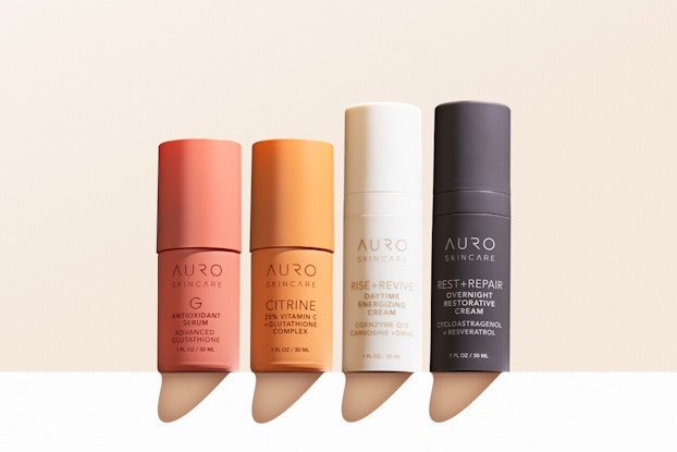 A line of Auro Wellness skincare products in cylindrical packages. From left to right: antioxidant serum in a rose pink tube, citrine in an orange tube, Rest + Revive daytime energizing cream in a white tube, and Rest and Repair overnight restorative cream in a dark gray tube.