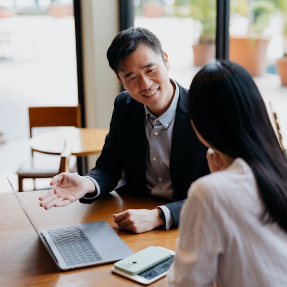 A sales representative and a prospective are seated at a table in a well-lit open space. The sales representative is gesturing to an open laptop sitting on the table while talking with the prospective customer.