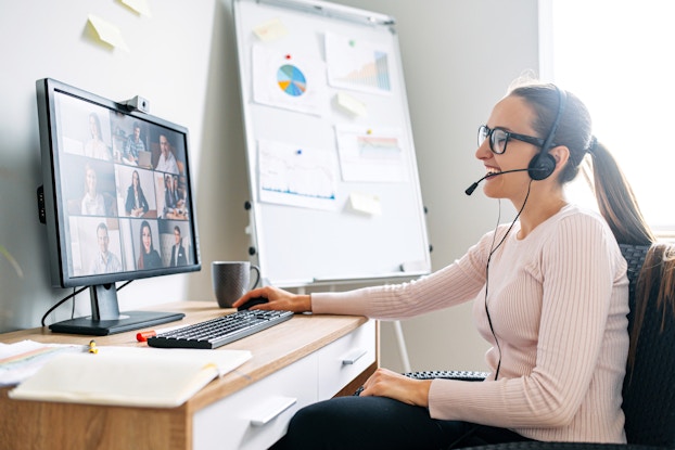 A young, smiling woman uses a computer to participate in a virtual meeting.