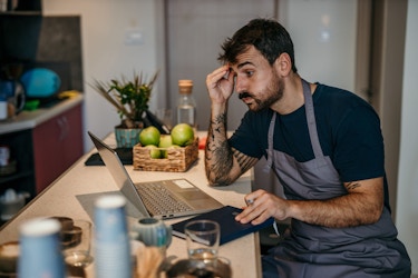 A concerned restaurant owner is seated at the restaurant bar looking at a laptop computer placed on the counter. He is reviewing his restaurant's finances.