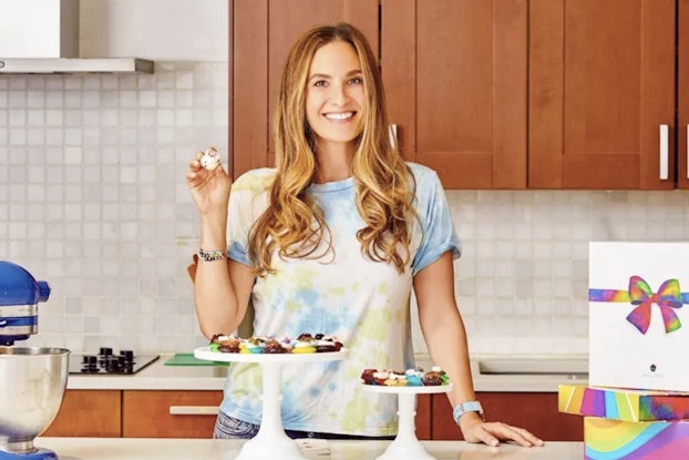A shot of Melissa Ben-Ishay, CEO and Co-founder of Baked by Melissa, standing at a kitchen counter, smiling, and holding one of her company's signature bite-size cupcakes. On the counter in front of Melissa are two cake stands, each holding several more tiny cupcakes. Melissa is a white woman with long, wavy blonde hair. She wears a yellow-and-blue pastel tie-dye T-shirt.