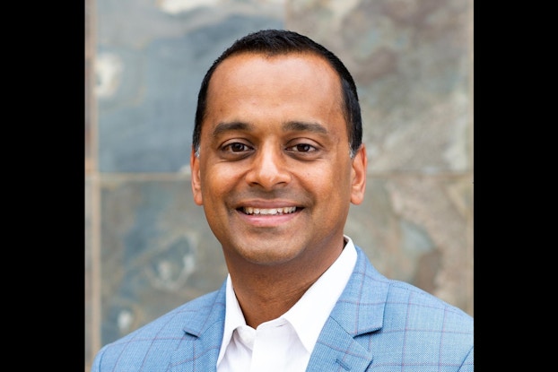 A close-up of Sanjay Khetan, Chief Financial Officer at Baked by Melissa. Sanjay is a South Asian man with short dark hair and brown eyes. He wears a white collared shirt and blue windowpane-checked blazer, and he smiles for the camera.