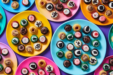An arrangement of several multicolored plates of bite-sized cupcakes, seen from above. Each plate holds at least a dozen small cupcakes, all in different flavors and colors, some topped with sprinkles or chocolate chips and some simply iced with white or brown icing.