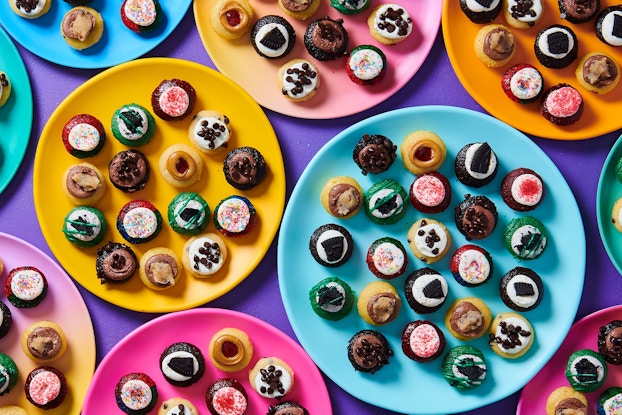 An arrangement of several multicolored plates of bite-sized cupcakes, seen from above. Each plate holds at least a dozen small cupcakes, all in different flavors and colors, some topped with sprinkles or chocolate chips and some simply iced with white or brown icing.