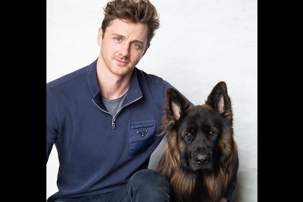 A two-shot of a Ordo Founder Barty Walsh and a dog. Walsh is a young man with brown hair and blue eyes. He sits with one arm around the dog, which has long black-and-brown fur and pointed ears.