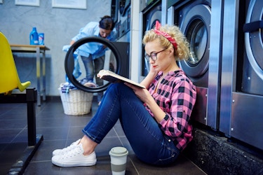  A woman sis on the floor of a laundromat reading a book while she waits for her laundry. Pictured next to her is a cup of coffee. 