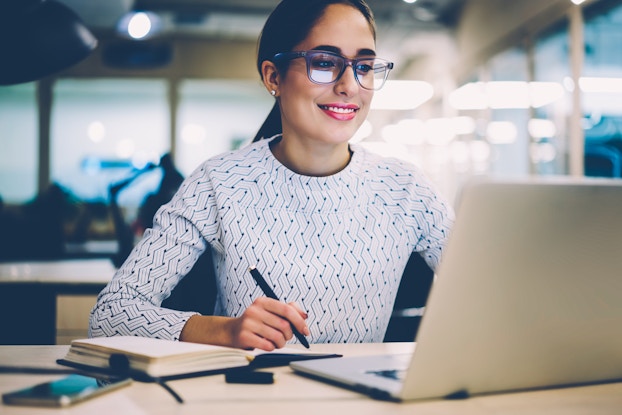  Young business woman is pleased with information she sees on her laptop and takes notes.