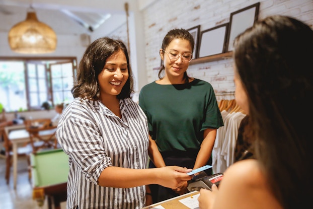  Two young women are in a small cafe. They are both smiling while one of them hold her debit card to a card reader to pay for her order.