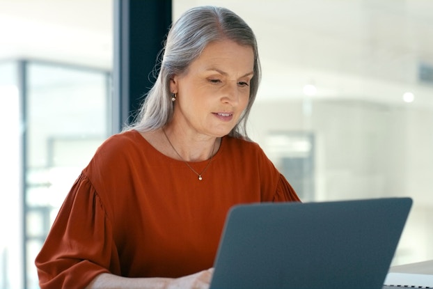A woman seated at a table types on a laptop computer. Behind her in the background is an office setting with office cubicles.