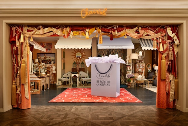 The entrance to Chairish's pop-up emporium at Bergdorf Goodman. The wide entrance is bordered with red curtain twisted with gold ribbon and large gold tassels. Through the entrance can be seen several mini-rooms displaying arrangements of vintage furniture. In the center of the room is a massive, person-sized white shopping bag, filled with white tissue paper and labeled with both the Chairish and Bergdorf Goodman logos.