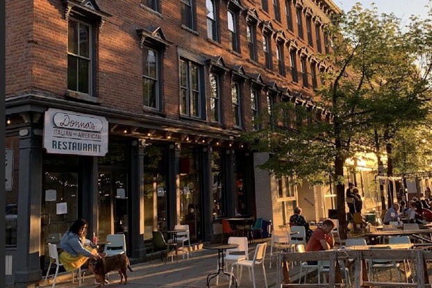 Outdoor seating outside a restaurant hosts customers with a tree and brick building in the background.