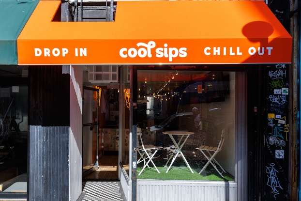 The exterior of a Cool Sips location in New York City. The shop has a glass front under an orange awning. The awning is printed with Cool Sips' logo between the phrases "DROP IN" and "CHILL OUT." Through the glass can be seen a slightly elevated seating area with two chairs and a small table on top of fake grass.