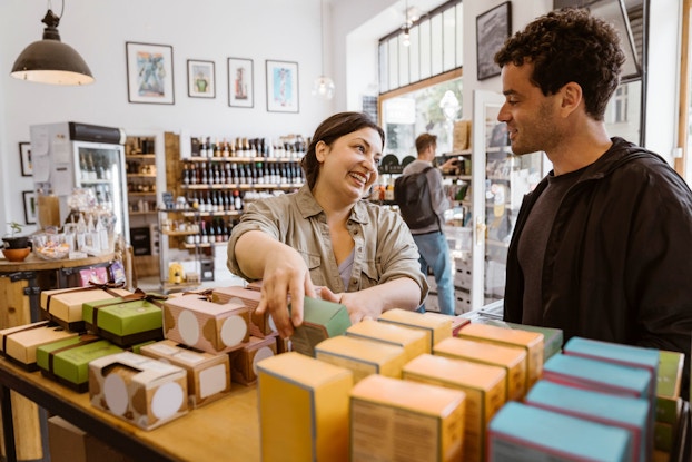 A smiling woman business owner talks to a male customer while holding up a small container.