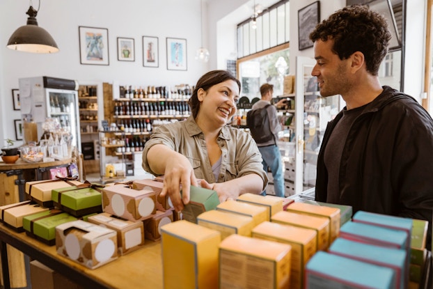  A smiling woman business owner talks to a male customer while holding up a small container.