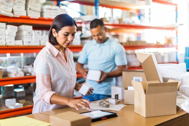 A woman shipping manager uses a tablet to prepare an item for shipment. Behind her is a man who is selecting an item from inventory to put in the shipment.