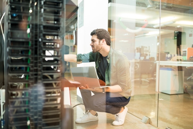 Network administrator with notebook computer sitting in data center room and working with networking device on rack cabinet.