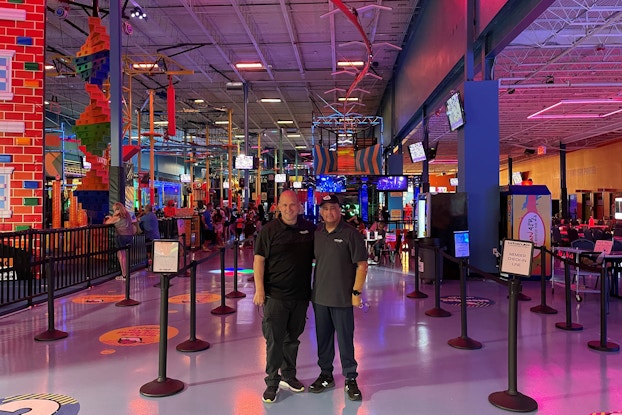  Two men stand next to each other inside an entertainment facility with crowd-control barriers and neon lights