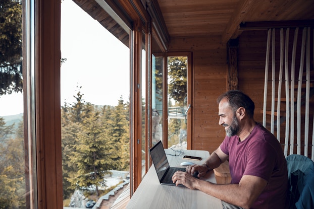 Close up of a mid-adult man working on a laptop from his cabin in the woods