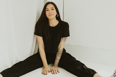 A picture of Dinah Chapman, Founder of Little Trouble, sitting on the floor in an all-white room. Dinah has long black hair and a sleeve tattoo on her left forearm. She sits with her legs spread in a V and wears a black jumpsuit and hoop earrings.
