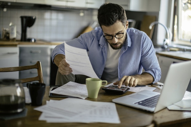 Man sits at kitchen table with laptop, papers, and calculator. He calculates information while drinking coffee.