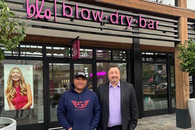 Don Padilla and Eric Abbe stand in front of their business, Blo Blow Dry Bar, a glass-fronted salon with its logo in pink above the entrance.