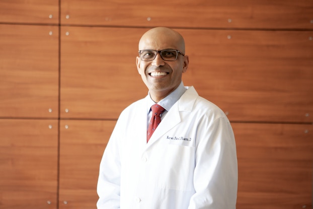 Dr. Nayan Patel, Founder and CEO of Auro Wellness, stands in front of a wood-paneled wall with a smile. Dr. Patel has a clean-shaven head and wears glasses and a lab coat embroidered with his name.