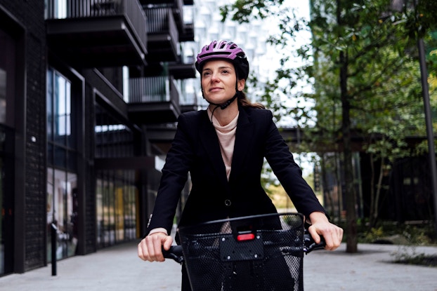  A businesswoman rides an electric bicycle in a city. She is wearing a purple bicycle helmet. Behind her is a street and an apartment building.