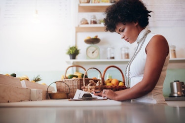  A woman business owner stands in a bright, modern kitchen. She has papers before her. She is reviewing and filling out a form. 