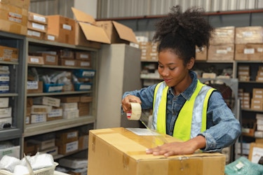  A young worker in a shipping warehouse prepares an order for shipment. Behind her are shelves stacked with boxes and various shipping materials. 