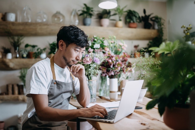 Male florist, owner of small business flower shop, working on laptop over counter against flowers and plants.