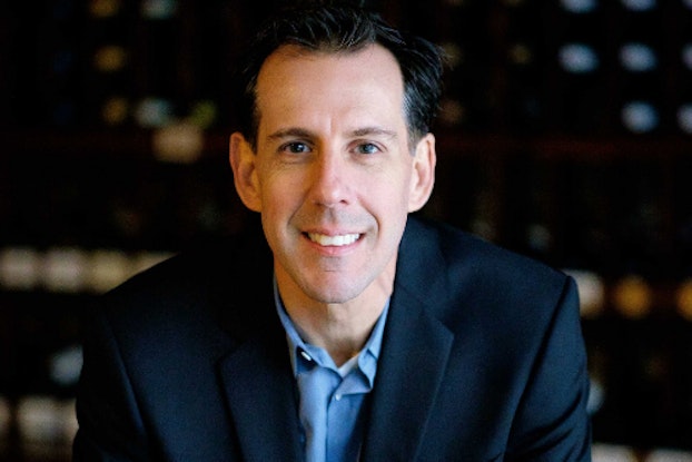 A shot of Eric Gagnon, President and Co-Founder of We Sell Restaurants, sitting with his hands folded and smiling. Gagnon is a white man with dark hair. He's wearing a dark blue blazer and light blue button-up shirt.