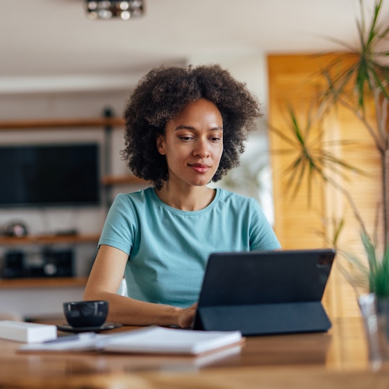 Woman works on laptop in home office with coffee mug and notebook on her desk.